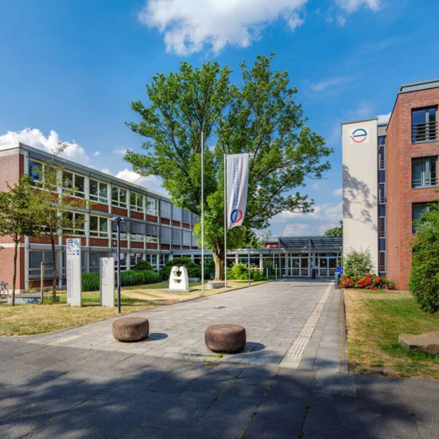 Haupteingang der EvH Bochum: Auf der rechten und linken Seite stehen große braun gepflasterte Hochschulgebäude mit vielen Fenstern. In der Mitte steht ein großer grüner Baum und eine Flagge mit dem rot-blauem Logo der Hochschule. Links und rechts neben den Gebäuden gibt es grüne Wiesen und einen Fahrradständer. Links vor der Gebäude steht eine weiße kleine Skulptur.