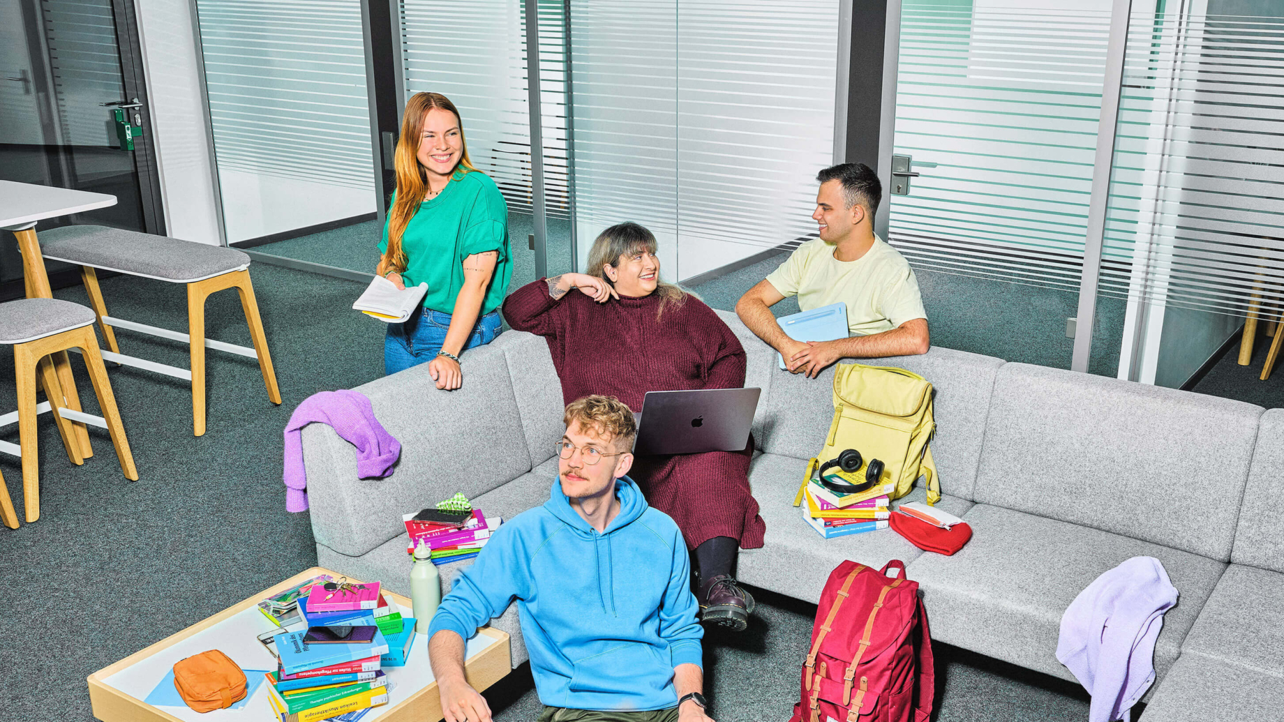 Das Bild zeigt 4 Studierende im Selbstlernzentrum. Im Fokus steht eine graue Eck-Couch auf der eine Frau in einem roten Kleid sitzt. Sie hat einen Laptop auf den Knien und unterhält sich mit einem Studenten, der rechts hinter der Couch kniet. Er trägt ein gelbes T-Shirt und hält ein Tablet in den Händen. Links von der Frau im roten Kleid steht eine Frau mit einem Buch in der Hand. Vor der Couch auf dem Boden sitzt ein Mann in hellblauem Hoodie mit einem Laptop auf dem Schoß. Sein Arm ist auf einen Beistelltisch gestützt, auf dem viele bunte Bücher liegen.