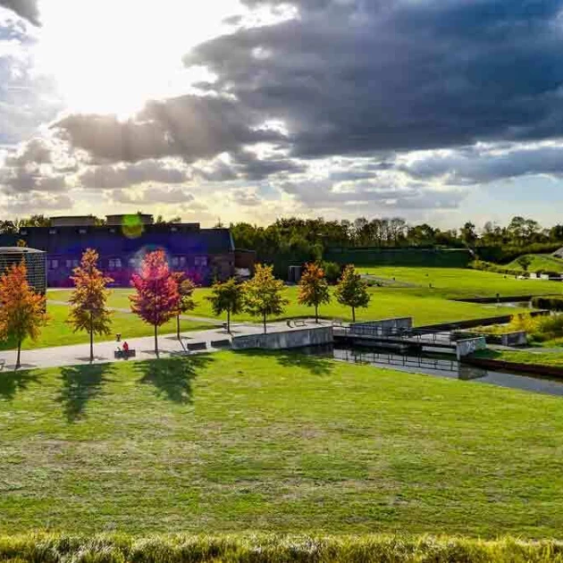 In einem Park stehen auf einer Wiese herbstliche Bäume mit bunten Blättern, ein Industriegebäude im Hintergrund und ein kleiner Wasserlauf. Die Sonne scheint durch Wolken hindurch.