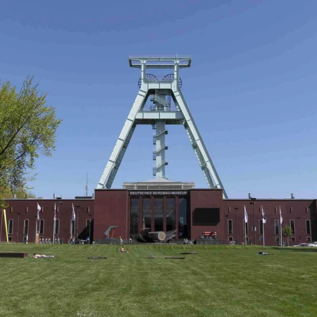 Ein großes braunes Gebäude steht auf einer grünen Wiese, es ist das Bergbaumuseum in Bochum. Darüber ist ein sehr großes und hellblaues Bergbaugerüst, mittig, platziert. Vor dem Museum steht ein großer grüner Baum.