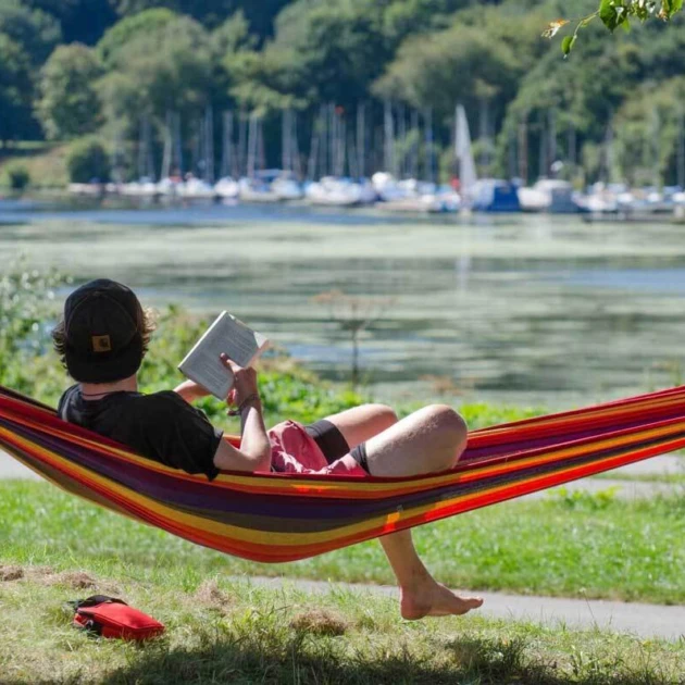 Ein Mann in dunklem T-Shirt und roter Badehose liegt in einer bunt gestreiften Hängematte. Er hält ein Buch in der Hand und hat den Kopf von der Kamera abgewandt. Er schaut auf einen großen See mit kleinen Booten und Bäumen im Hintergrund.