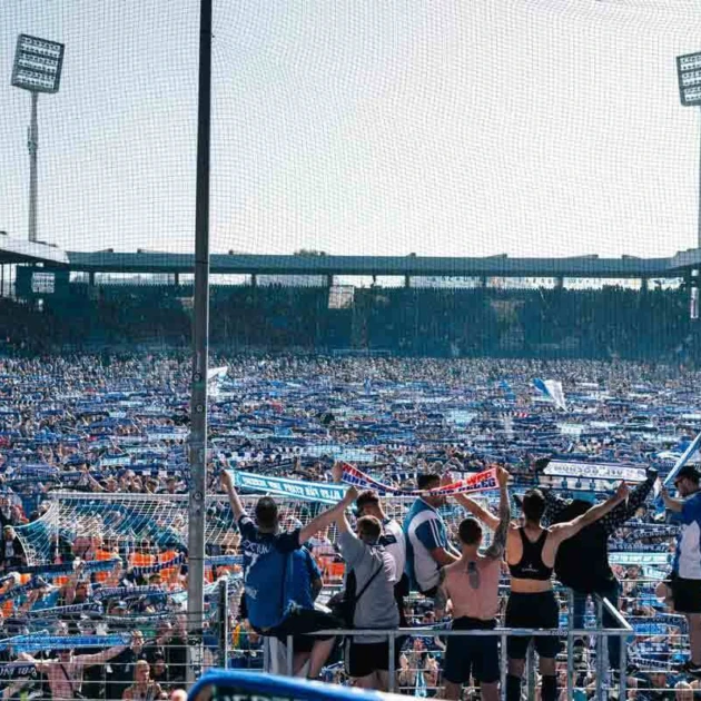 Ein Fußballstadion ist mit Menschen gefüllt, die dicht aneinander stehen und blaue Fahnen und Schals in die Luft halten. Auf einer Tribüne stehen mehrere Fußballspieler mit dem Rücken zur Kamera und jubeln in die Menge.