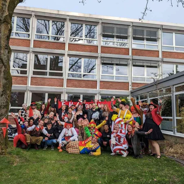 Eine Gruppe von Studierenden hat sich als Zirkuspersonen verkleidet und steht von dem Hochschulgebäude auf einer Wiese. Sie jubeln fröhlich in die Kamera.