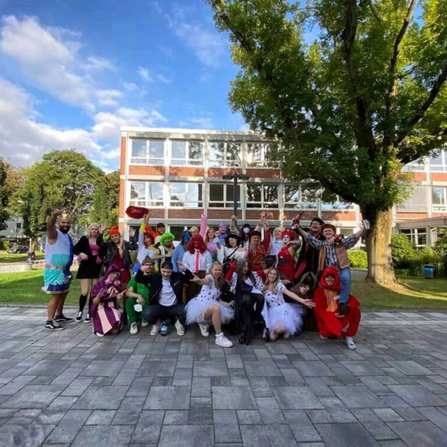 Eine Gruppe als Disney-Figuren verkleidete Studierende steht vor einer grünen Wiese, einem Baum sowie dem Hochschulgebäude der EvH Bochum. Sie jubeln fröhlich in die Kamera.