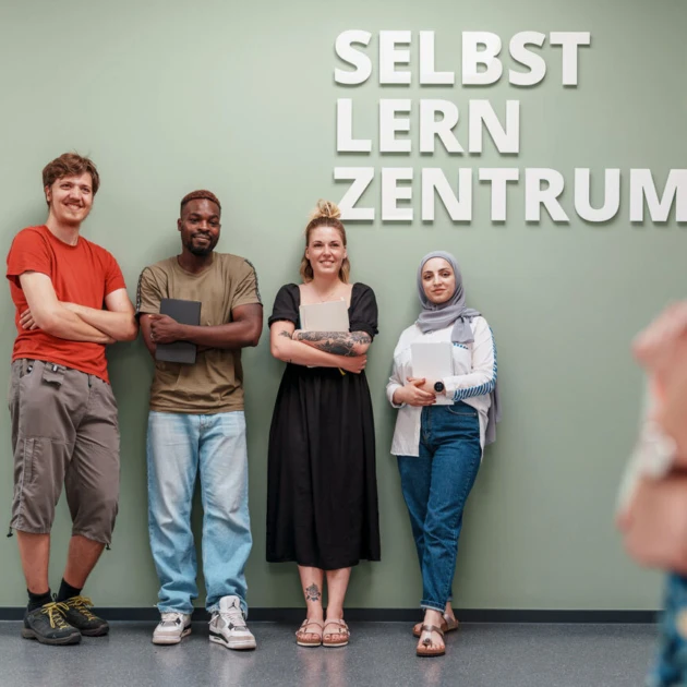 An einer grünen Wand lehnen 4 Studierende nebeneinander und lächeln in die Kamera. Der Mann links trägt ein rotes T-Shirt, eine 3/4 Hose und hat die Arme locker vor der Brust verschränkt. Der Mann rechts neben ihm trägt ein olivgrünes T-Shirt und eine hellblaue Jeans. Er hält ein Tablet in der Hand. Die Frau rechts neben ihm trägt ein schwarzes sommerliches Kleid. Sie hat einen Block in der Hand. rechts neben ihr steht eine Frau mit weißer Bluse, Jeanshose und Kopftuch. Auch sie hat einen Block in der Hand. Oben rechts auf der Wand steht in weißer Schrift "Selbstlernzentrum".