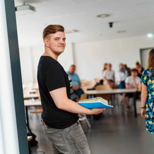 Ein Student steht in der Tür eines Seminarraums und hat schaut über die Schulter in die Kamera. Er trägt ein schwarzes T-Shirt, eine graue Jeans und hält einen Block und ein dickes Buch auf dem Arm. Im Hintergrund sitzen weitere Studierende auf Tischen und Stühlen.