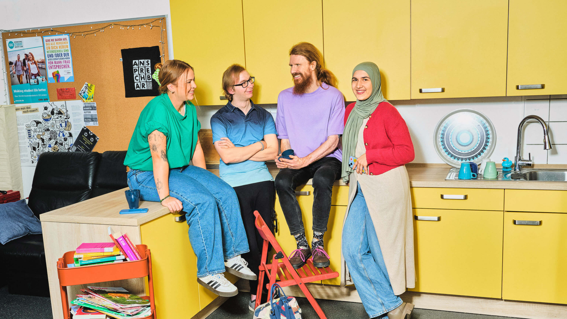 Eine Gruppe von 4 Studierenden steht in einer gelben Teeküche. Eine Frau in rotem Cardigan lächelnd auffordernd in die Kamera. Sie hält ein Handy in der Hand und trägt ein Kopftuch. Links neben ihr sitzt ein Mann mit roten Locken auf der Küchenzeile. Er trägt ein lila T-Shirt, hat die Haare zu einem Zopf gebunden und einen Vollbart. Er blickt auf den Mann links neben sich und hat die Beine vor sich auf einem roten Klappstuhl aufgestellt. Der Mann neben ihm hat blonde Haare, eine Brille und trägt ein T-Shirt mit blauem Farbverlauf. Die Arme hat er verschränkt und er blickt lächelnd nach rechts. Links neben ihm sitzt eine Frau mit grünem T-Shirt auf der Küchenzeile. Sie trägt ihre roten Haaren mit einer Klammer zusammengebunden und stützt sich mit den Armen auf die Arbeitsplatte auf. Im Hintergrund ist eine Pinnwand, ein schwarzes Sofa und ein Beistelltisch mit bunten Zeitschriften zu erkennen.