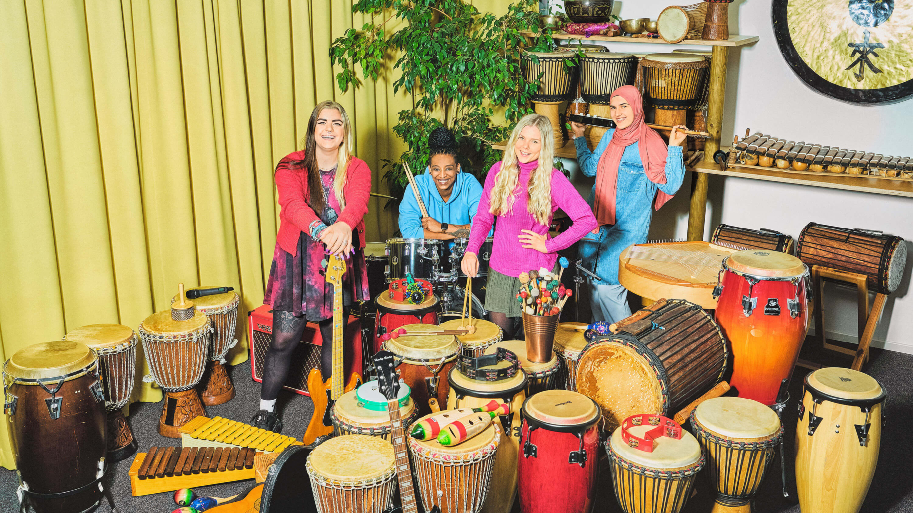 4 Studentinnen stehen in einem Musikraum zwischen verschiedenen Instrumenten. Die Studentin auf der linken Seite hat schwarz-blonde Haaren, trägt eine rote Strickjacke über einem rot-schwarzen Kleid hat die Hände auf eine Bassgitarre gestützt. Neben ihr sitzt eine Studentin an einem Schlagzeug. Sie trägt einen hellblauen Hoodie und ihre Braids in einem Dutt. Vor ihr steht eine Studentin mit blonden lockigen Haaren und einem pinken Pullover, die ihre Schläger auf die Trommel vor sich stützt. Rechts hinten hält eine Studentin eine Bass-Gitarre hinter ihrem Kopf. Sie trägt ein rosanes Kopftuch und ein hellblaues Kleid. Der Raum ist voll mit unterschiedlichen Musikinstrumenten, wie Gitarren und Trommeln unterschiedlicher Größe. Im Hintergrund steht eine große grüne Zimmerpflanze.