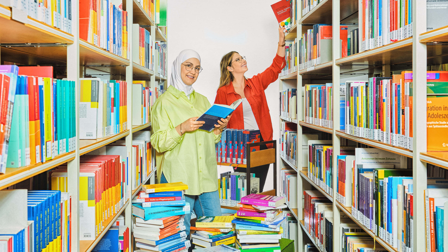 2 Studentinnen stehen in einem Gang aus zwei Bücherregalen, um sie herum sind bunte Bücher aufgetürmt. Eine Studentin hält ein Buch in der Hand und lächelt in die Kamera. Sie trägt eine gelbe Bluse, blaue Jeans, ein weißes Kopftuch und eine Brille. Hinter ihr zieht eine andere Studentin ein Buch aus dem Regal. Sie trägt eine rote Bluse offen über einem weißen T-Shirt., schwarze Jeans und eine Brille. Ihre blonden Haare hat sie hinter die Ohren gestrichen.