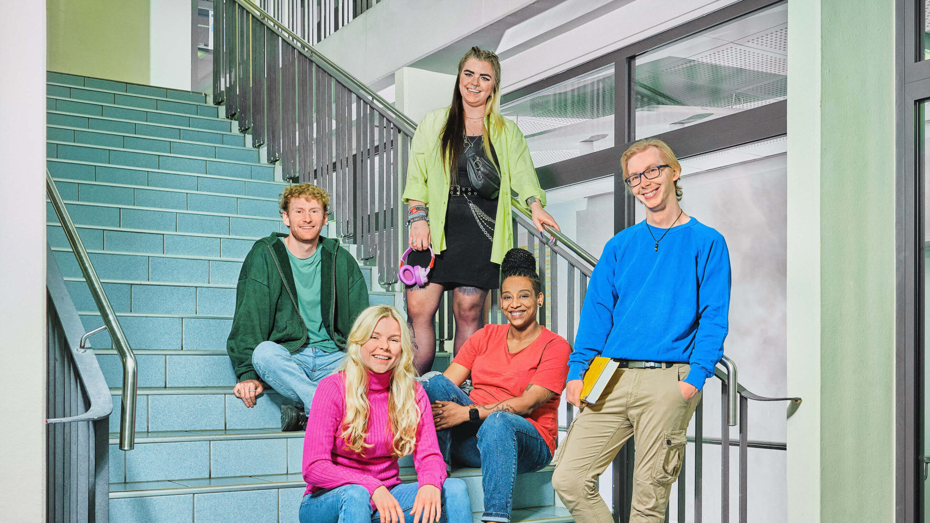 Eine Gruppe von 5 Studierenden sitzt und steht auf einer großen blauen Treppe. Personenbeschreibung von links vorne nach hinten: Eine Studentin sitzt auf einer Treppenstufe. Sie hat die Arme locker auf die Knie aufgestützt. Sie trägt einen pinken Pulli, blaue Jeans und schwarze Stiefel. Ihre welligen blonden Haare sind offen. Rechts neben ihr steht ein Student mit einem Buch unterm Arm. Er hat sich ans Treppengeländer angelehnt und hat die andere Hand in der Hosentasche. Er trägt einen blauen Pulli, eine beige Hose, eine schwarze Kette mit Anhänger, eine Brille und hat blonde Haare. Hinter ihm sitzt eine Studentin ans Geländer gelehnt. Sie trägt ein rotes Tshirt, blaue Jeans und ihre Braids in einem Dutt. Links oben sitzt ein Student im Schneidersitz auf einer Treppenstufe, die Hände hat er auf die Stufe gestützt. Er trägt einen grünen Pulli offen über einem grünen Tshirt und hellblaue Jeans. Er hat rote lockige Haare. Rechts neben ihm steht eine Studentin ans Treppengeländer gelehnt, die stützt sich mit der einen Hand aufs Geländer und hält in der anderen pinke Kopfhörer. Sie trägt eine gelbe Bluse offen über einem schwarzen Kleid und eine Bauchtasche quer über dem Oberkörper. Ihre Haare sind schwarz und blond gefärbt und zu 2 Zöpfen gebunden.