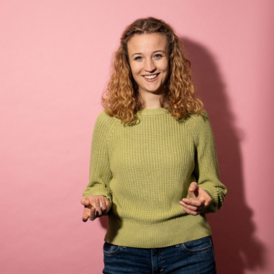 Eine junge Frau lächelt freundlich in die Kamera. Sie hat blonde offene, lockige Haare und trägt einen hellgrünen Strickpullover mit einer blauen Jeans. Sie steht vor einem rosa Hintergrund.