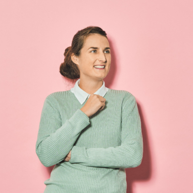 Eine junge Frau lächelt freundlich. Sie trägt ihre braunen Haare in einem Dutt. Einen Am hat sie verschränkt, der andere ist angewinkelt. Sie trägt unter einem pastellgrünen Pullover ein weißes Hemd mit Kragen. Die Jeans der Frau ist hellblau. Der Hintergrund vor dem die Frau steht ist rosa.