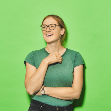 Eine junge Frau lächelt freundlich an der Kamera vorbei. Sie hat blonde Haare zu einem Zopf gebunden und trägt eine Brille. Das T-Shirt ist dunkelgrün und die Jeans ist schwarz. Sie trägt eine Armbanduhr und verschränkt ihren einen Arm unter dem anderen. Der andere Arm liegt auf dem Brustkorb auf. Die Frau steht vor einem grünen Hintergrund.